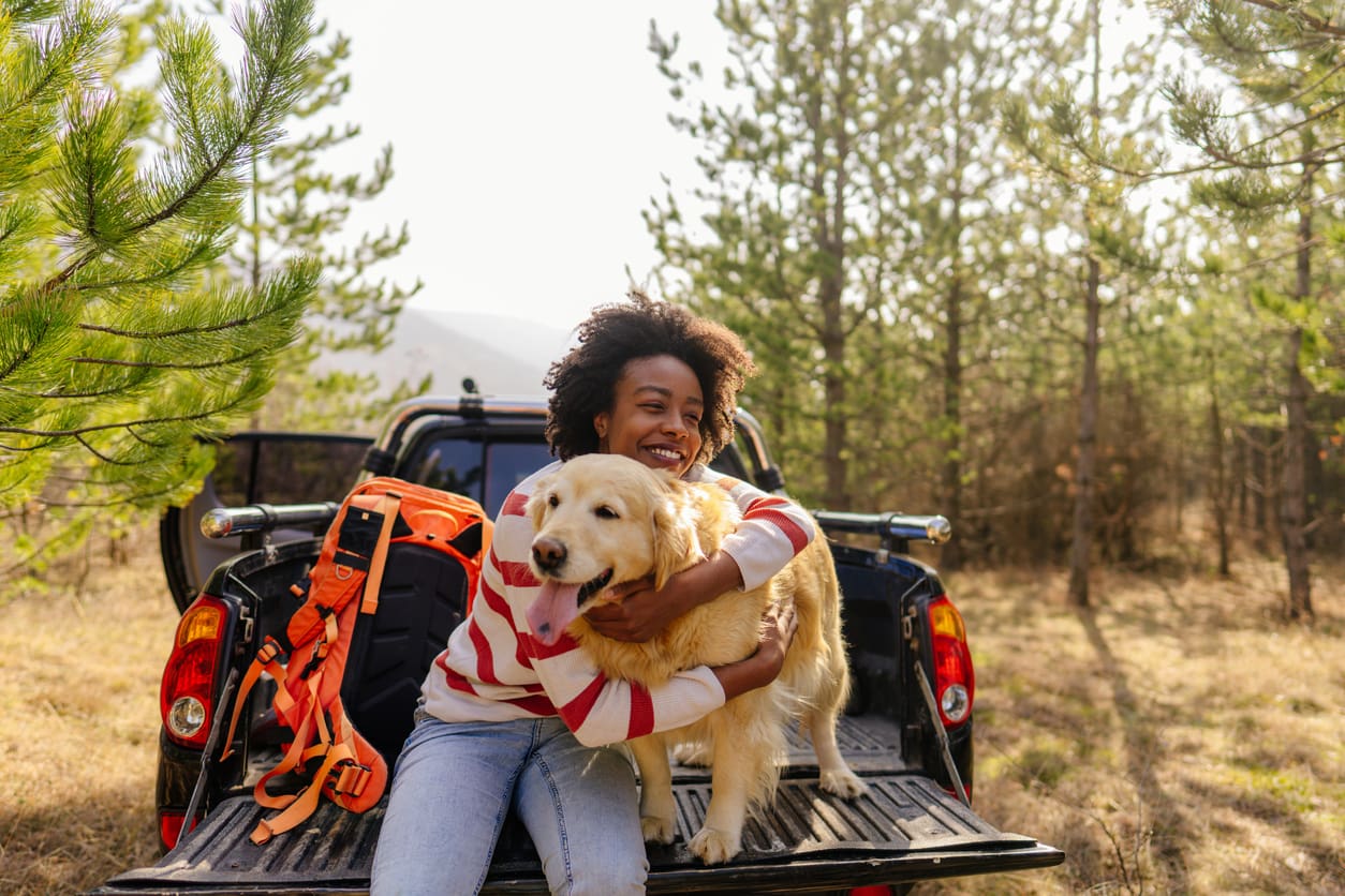 A smiling child hugs a golden retriever while sitting on the open tailgate of a pickup truck in a sunny forest, showcasing the joy of pet travel safety. An orange backpack is nearby, and trees surround them in the background.