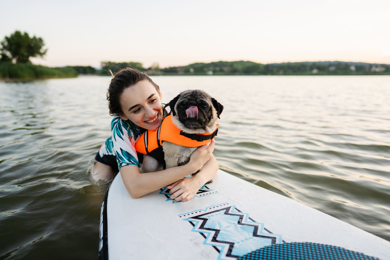 A smiling woman in the water hugs a pug in an orange life jacket while sitting on a paddleboard in a lake, highlighting dog beach safety with trees and hills in the background.