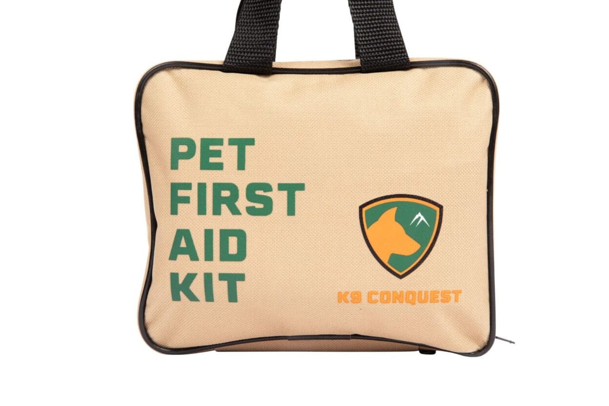 A beige pet first aid kit bag with black handles, green PET FIRST AID KIT text, and an orange-green shield logo featuring a dogs head and mountain—ideal for tackling trail hazards like foxtails during summer hiking dangers for dogs. Labeled K9 CONQUEST.